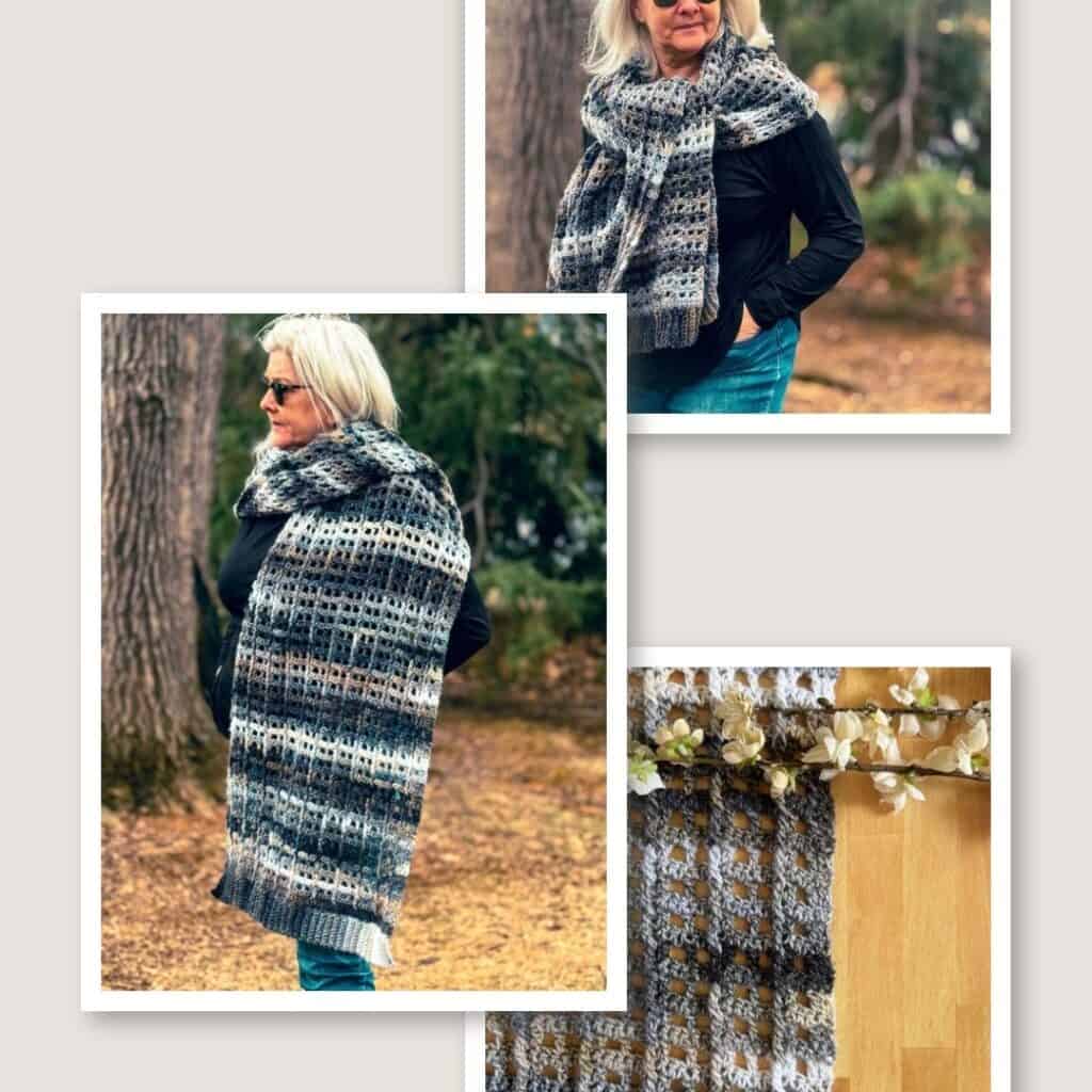 A woman with short white hair and sunglasses stands outdoors in a wooded area, wearing a black top and blue jeans. She is wrapped in a handmade granny stitch shawl featuring a textured, openwork stitch pattern with a gradient of earthy tones, including shades of gray, brown, and cream. The shawl drapes over her shoulders and extends down her back. The background consists of trees and fallen leaves, adding to the natural, cozy atmosphere.