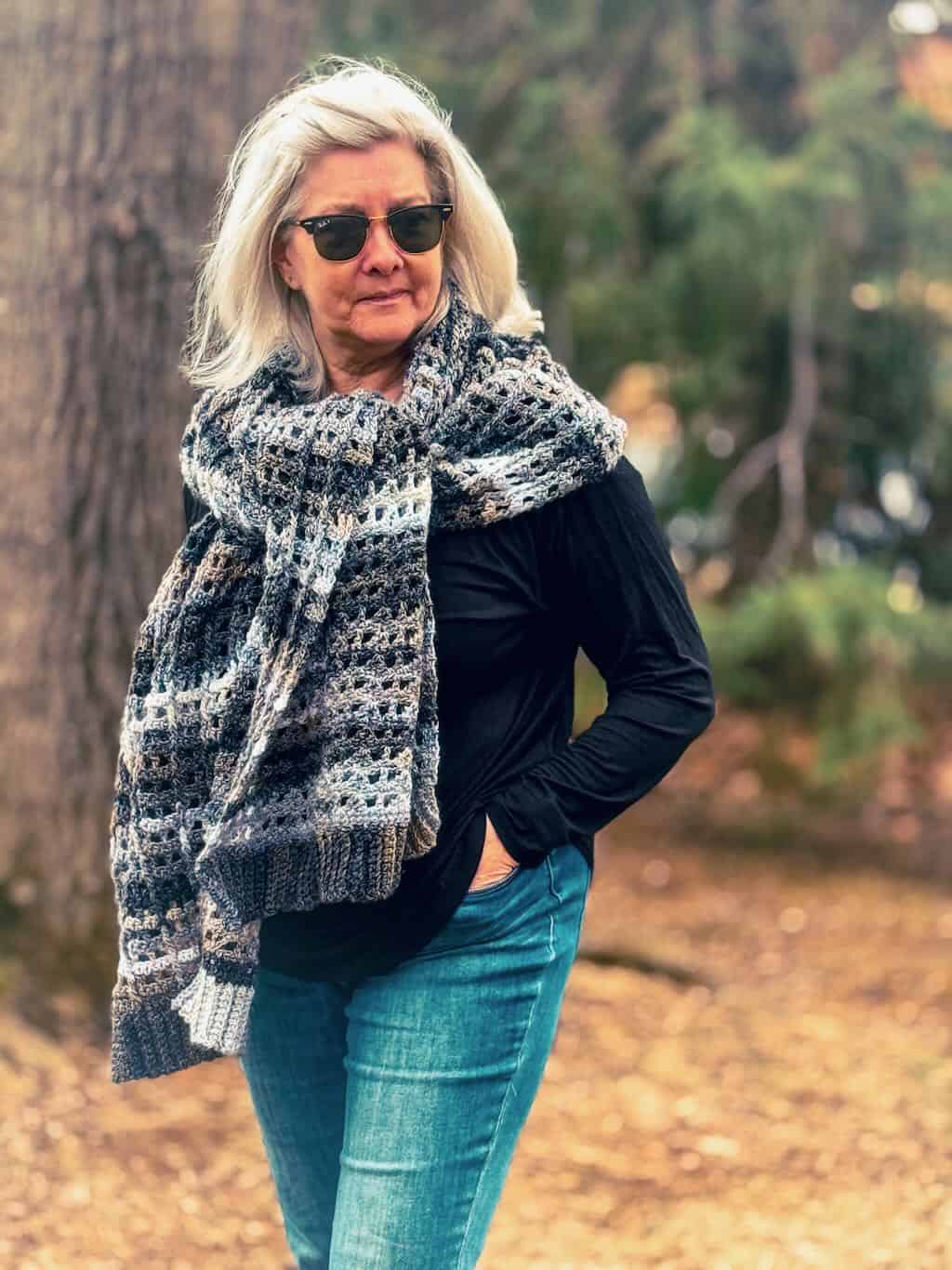 A woman with short white hair and sunglasses stands outdoors in a wooded area, wearing a black top and blue jeans. She is wrapped in a handmade granny stitch shawl featuring a textured, openwork stitch pattern with a gradient of earthy tones, including shades of gray, brown, and cream. The shawl drapes over her shoulders and extends down her back. The background consists of trees and fallen leaves, adding to the natural, cozy atmosphere.