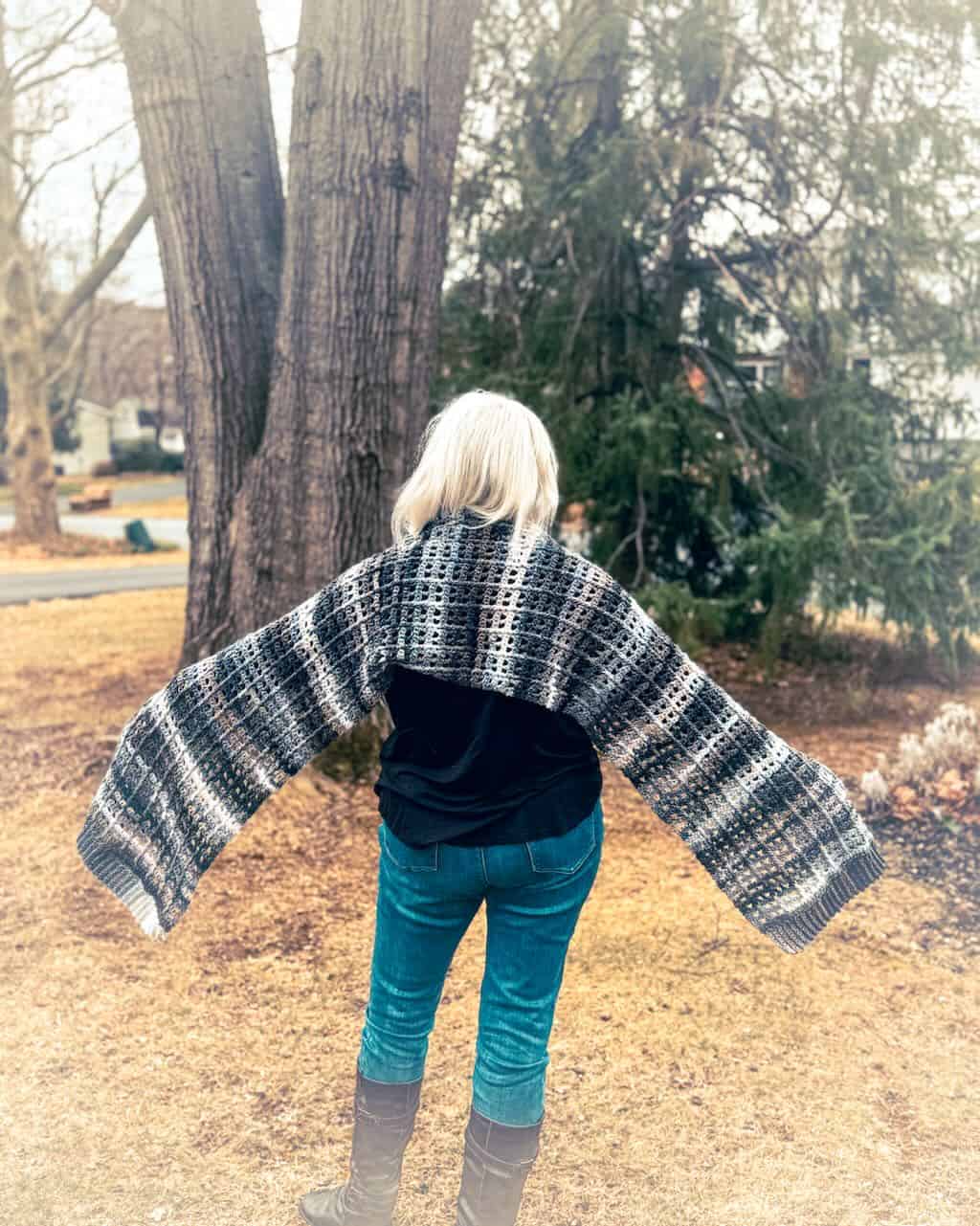 A woman with short white hair and sunglasses stands outdoors in a wooded area, wearing a black top and blue jeans. She is wrapped in a handmade granny stitch shawl featuring a textured, openwork stitch pattern with a gradient of earthy tones, including shades of gray, brown, and cream. The shawl drapes over her shoulders and extends down her back. The background consists of trees and fallen leaves, adding to the natural, cozy atmosphere.